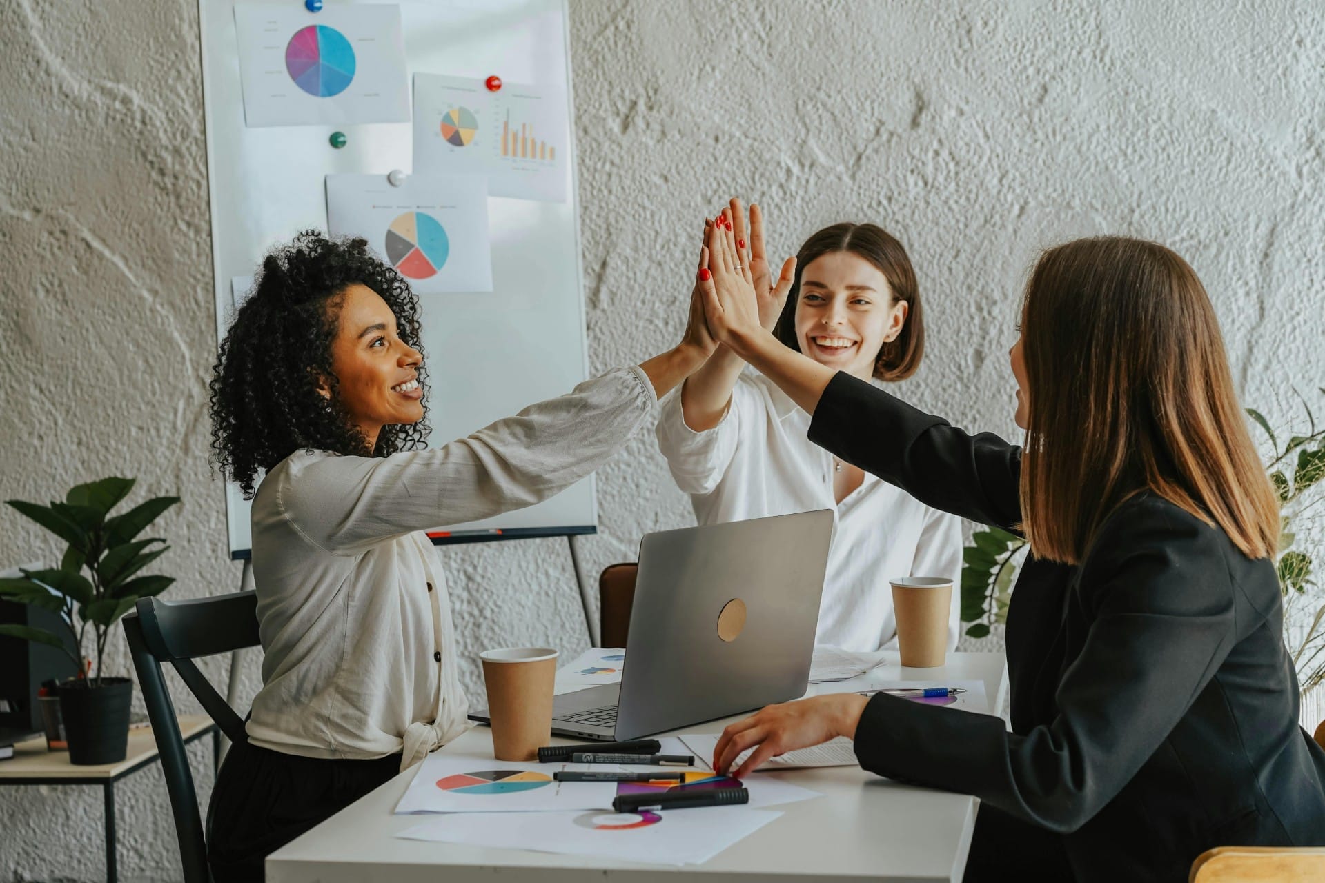 Three women in an office setting high-five around a table with a laptop, coffee cups, and charts. A whiteboard with graphs is in the background