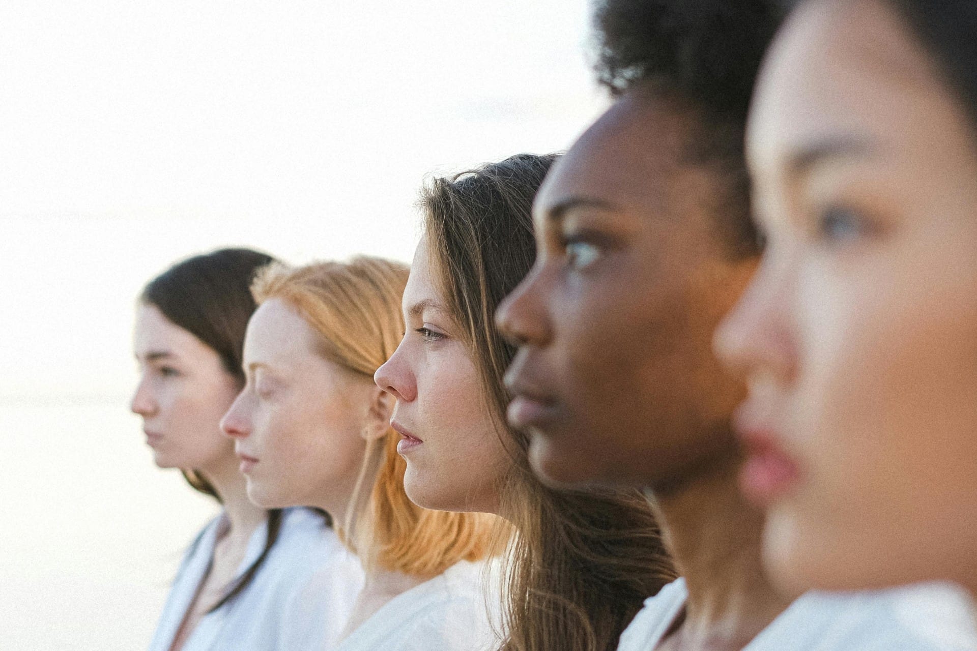 A group of five women of diverse backgrounds and appearances stand side by side, all facing the same direction against a light background.